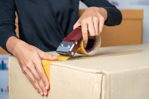 Close up of woman hands holding packing machine and sealing cardboard boxes stock photo