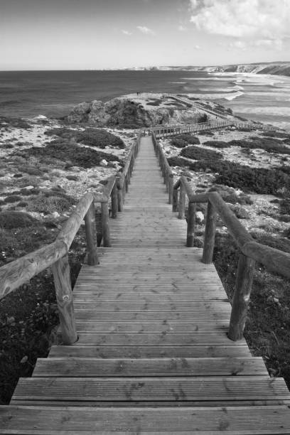 close up of wooden pathway with overhanging view on atlantic coast by the beach of bordeira from cliff ledge with breaking waves in black and white, popular surf spot destination in algarve, portugal stock photo