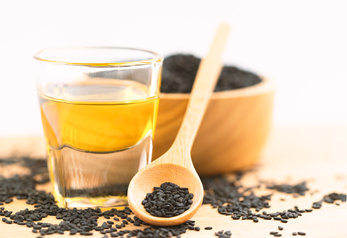 Close up organic black sesame oil in small glass with sesame seeds in wooden spoon and bowl, some black sesame seeds spread on wooden table, warm natural bright light, white background.