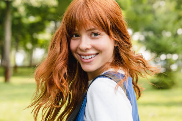 Close up portrait of a young caucasian woman girl teenager student schoolgirl with ginger red hair and toothy smile walking in park forest outdoors looking at camera. stock photo