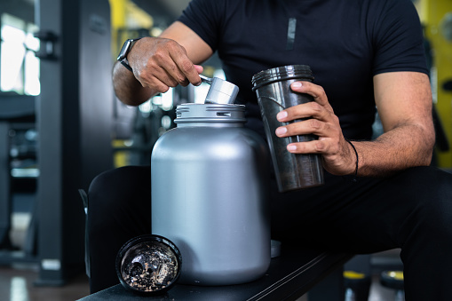 close up shot of bodybuilder hands taking protein powder and mixing with water on bottle by shaking at gym - concept of muscular gain, nutritional supplement and wellness