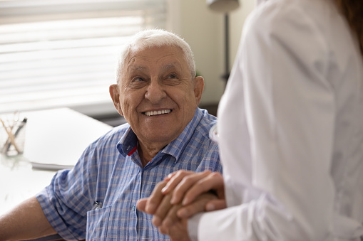 Close up smiling mature man and female caregiver wearing white uniform holding hands, doctor nurse comforting and supporting senior patient at meeting in hospital, expressing empathy and care