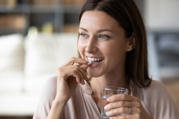 Close up smiling woman taking pill, holding water glass Close up smiling woman taking white round pill, holding water glass in hand, happy young female taking supplement, daily vitamins for hair and skin, natural beauty, healthy lifestyle vitamin stock pictures, royalty-free photos & images