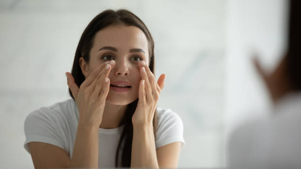 Close up smiling young woman doing facial massage, applying cream Close up smiling young woman wearing white t-shirt doing facial massage, applying moisturizing cream on under eye skin, looking in mirror, standing in bathroom, enjoying skincare procedure eye cream stock pictures, royalty-free photos & images