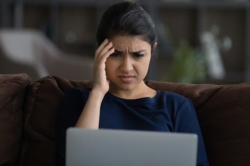 Close up unhappy Indian woman looking at laptop screen, reading bad news in message or social network, sitting on couch at home, worried businesswoman freelancer received dismissal notification