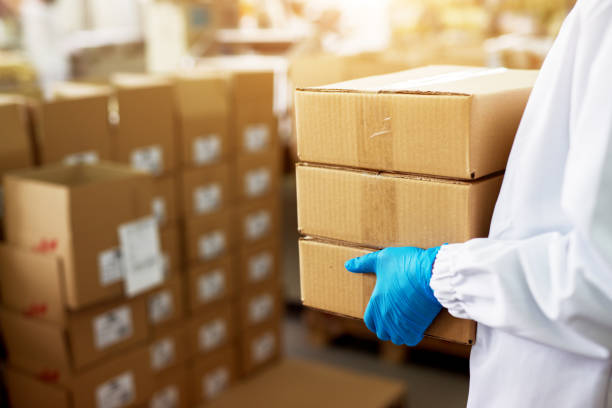 Close up view of a dedicated worker carrying a stack of duck taped brown boxes in factory storage room while wearing sterile cloths and rubber gloves. Close up view of a dedicated worker carrying a stack of duck taped brown boxes in factory storage room while wearing sterile cloths and rubber gloves. packaging stock pictures, royalty-free photos & images
