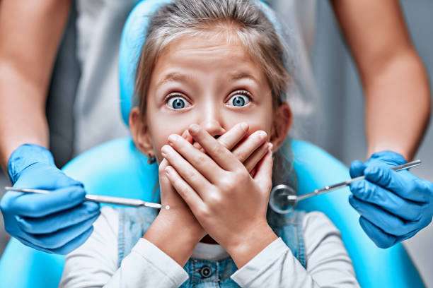 Close up view of a little girl looking scared and terrified screaming covering her mouth from the dentists with medical tools Close up view of a little girl looking scared and terrified screaming covering her mouth from the dentists with medical tools. Front view scared of dentist stock pictures, royalty-free photos & images