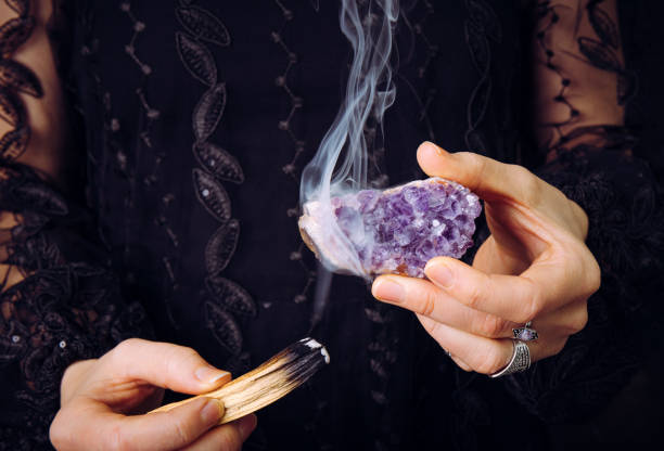 Close up view of woman in black lace dress, cleansing amethyst crystal cluster gemstone by smudging Palo Santo wood stick. Remove negative energy. stock photo