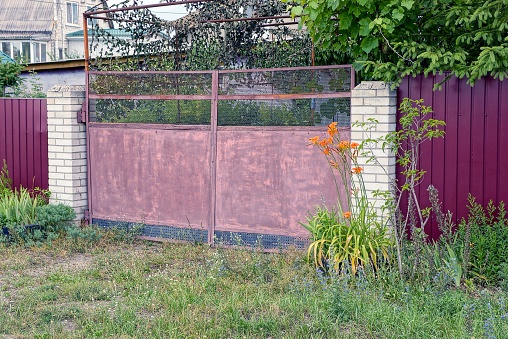 Closed Old Metal Gates Of Red Color And Part Of A Fence Overgrown