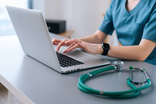 Close-up hands of unrecognizable female physician in medical uniform working typing on laptop keyboard sitting at desk stock photo