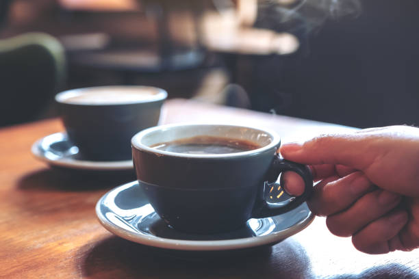Imagen de primer plano de una mano sosteniendo una taza azul de café caliente con humo en una mesa de madera en una taza de café y platillo fotografías e imágenes de stock