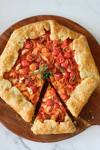 Stock photo showing elevated view of freshly cooked savoury galette, homemade, golden pastry crust, containing pesto, cherry tomato halves, crumbled feta cheese, sprinkled with rosemary leaves. Free form, rustic pie left to cool.