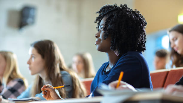 Close-up of a Beautiful Black Female Student Sitting Among Her Fellow Students in the Classroom, She's Writing in the Notebook and Listens to a Lecture.  lecture stock pictures, royalty-free photos & images