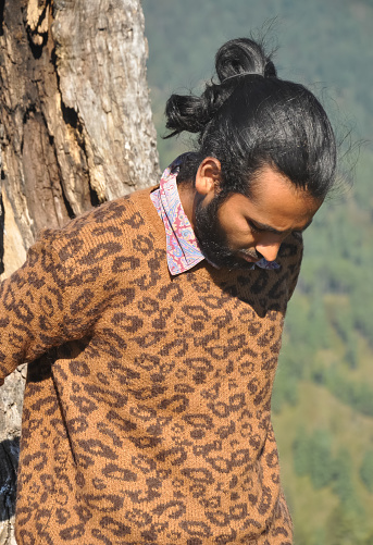 Closeup of a good looking Indian young men with hair bun looking down while leaning against tree