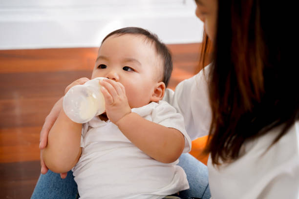 在家裡的客廳裡，亞洲母親在客廳裡用奶瓶給兒子餵奶。- 母性和配方餵養新生兒概念 - baby formula 個照片及圖片檔
