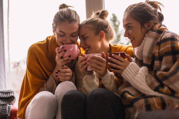 Close-up of happy close female friends sharing cozy time Close-up of three happy close female friends in woolen clothes sitting by the window while sharing hot beverage time. hygge stock pictures, royalty-free photos & images
