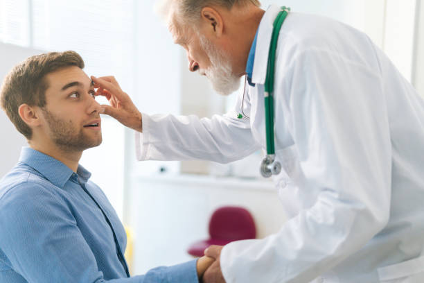 Close-up of mature adult male ophthalmologist examining eyes of male patient during checkup visit in clinic office. stock photo