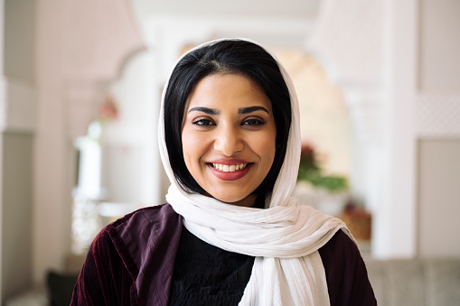 Head and shoulders portrait of woman with black hair wearing abaya, white headscarf, and smiling at camera in hotel lobby with Islamic architectural design.