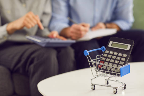 Close-up of shopping trolley and calculator with people calculating expenses in background stock photo