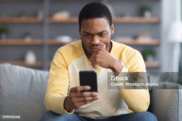 Closeup of thoughtful black guy sitting on couch, using smartphone