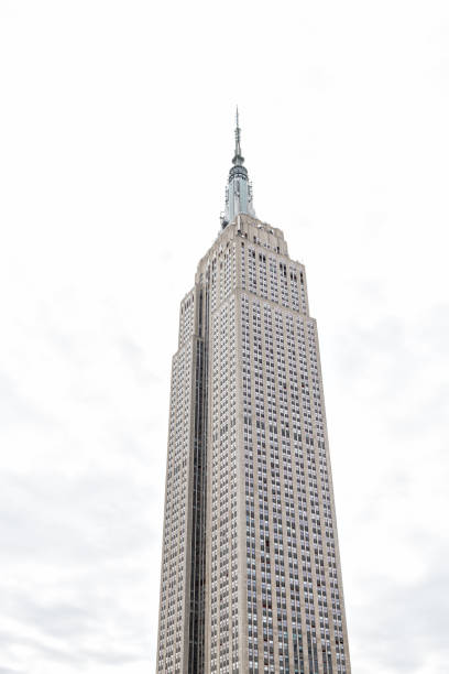 Closeup of top of empire state building isolated against cloudy white sky during day rooftop famous iconic building in NYC Herald Square Midtown, high tall spire stock photo