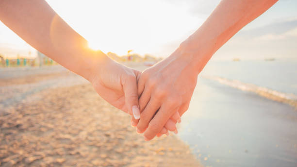 Close-up of two female holding hands against sunset - two girls walking on the beach hand by hand - same-sex lesbian couple and female friendship and love, celebration of Valentine's Day concept. stock photo
