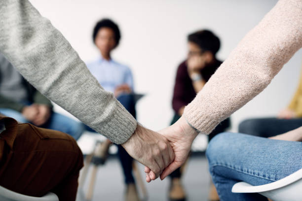 Close-up of two people holding hands during group therapy. Close-up of couple holding hands while attending psychotherapy with group of people. group therapy stock pictures, royalty-free photos & images