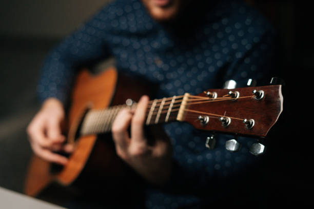 Close-up of unrecognizable guitarist singer male playing acoustic guitar sitting on armchair in dark living room, selective focus. stock photo