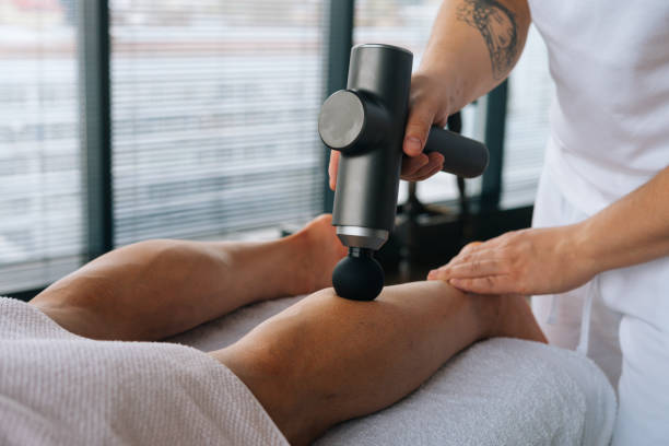 Close-up of unrecognizable professional male masseur massaging leg calf muscles using massage gun percussion tool of muscular athlete man, stock photo