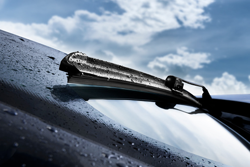 Close shot of a wet windscreen wiper blade wipping a car's window with a blue sky with clouds on the background