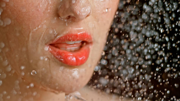 Close-up of woman being splashed with water Young woman's face being splashed with water against black background. orange lipstick stock pictures, royalty-free photos & images