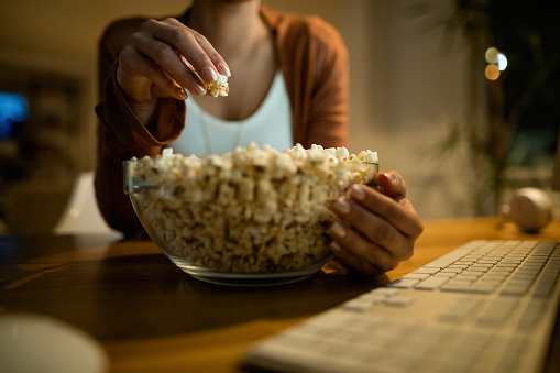 Close-up of unrecognizable woman eating popcorn while using computer at night at home.