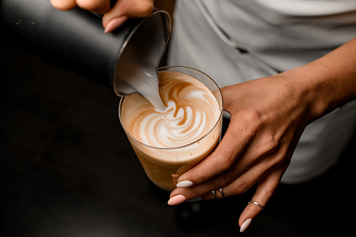 Close-up of woman who beautiful drawing pattern pouring milk into glass with latte in her hand