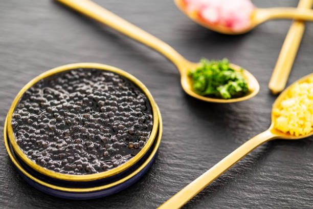 Closeup on high quality real black sturgeon caviar in a golden tin can aside golden spoons with pickles and herbs on a slate background. Side view. stock photo