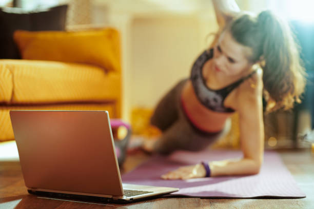 Closeup on laptop and woman in background doing cardio exercises Closeup on beige laptop and woman in background doing cardio exercises on fitness mat while watch fitness streaming on internet in the modern living room. personal trainer stock pictures, royalty-free photos & images