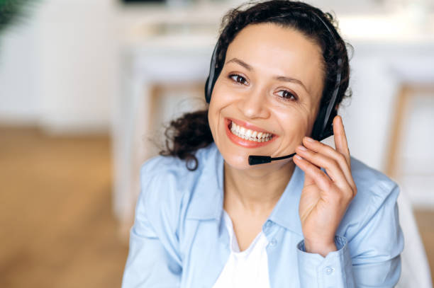 Close-up portrait of friendly successful mixed race adult woman, call center operator, agent, business person, wearing headphones, looking at the camera, smiling friendly stock photo