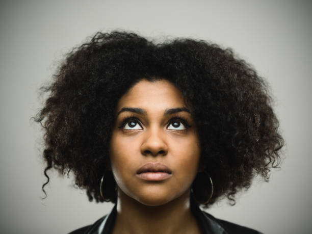 Close-up portrait of real young black woman looking up Close-up portrait of real young black woman looking up against gray background. Beautiful woman with curly hair. Studio photography from a DSLR camera. Sharp focus on eyes. emotional series stock pictures, royalty-free photos & images