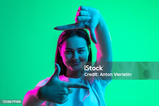 istock Close-up portrait of young pretty smiling caucasian girl showing frame gesture isolated on green background in neon light. 1328679188