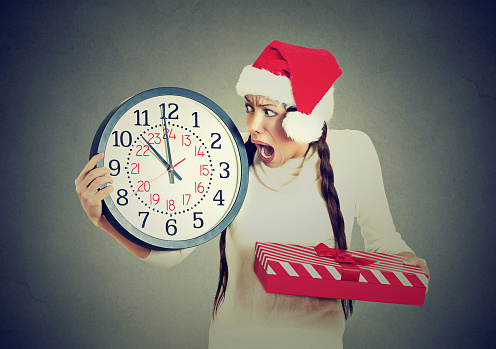 Closeup portrait worried stressed in a hurry young woman wearing red santa claus hat holding clock gift box