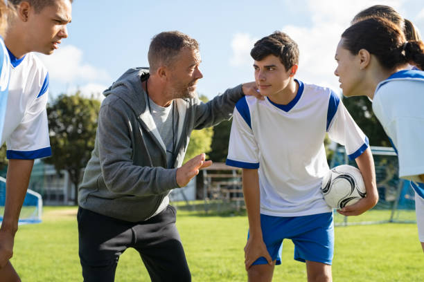 Coach talking to young soccer team before the match Mature coach teaching strategy to high school team on football field. Young soccer players standing together united and listening coach motivational speech. Coach giving team advise before school match. coach stock pictures, royalty-free photos & images