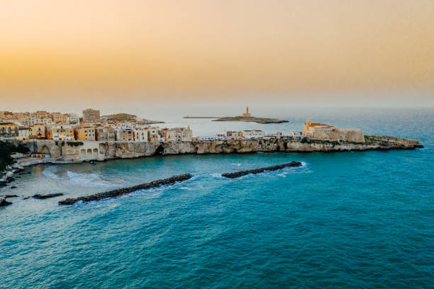 Coastal old town on peninsula at sunset, Vieste, Puglia, Italy Aerial view of old buildings in a scenic old town on a small peninsula on coastline under a clear orange sky during sunset, Vieste, Puglia, Italy puglia stock pictures, royalty-free photos & images