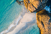 istock Coastline aerial photograph of aquamarine ocean and man walking along white sandbar beach 1299198919
