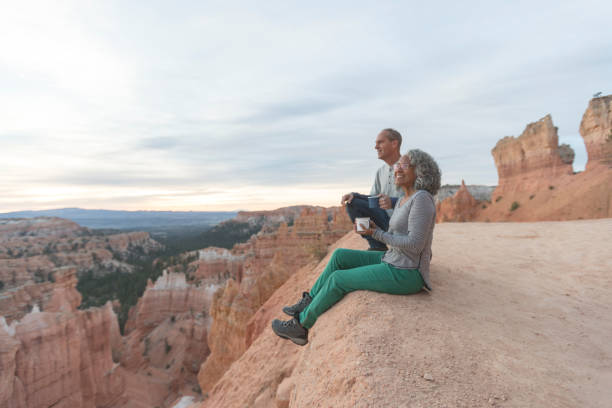 Coffee on a Cliff An older couple take a break from hiking to enjoy the view at Utah outlook overlooking a canyon. They are sitting on the cliff's edge and soaking in the scenery. The mountains and canyon are in front of them. bryce canyon national park stock pictures, royalty-free photos & images