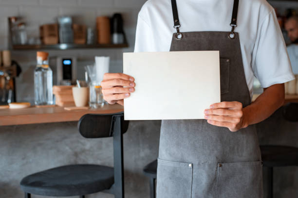 Coffee shop owner in apron holding blank sheet in hands while standing in coffeehouse stock photo