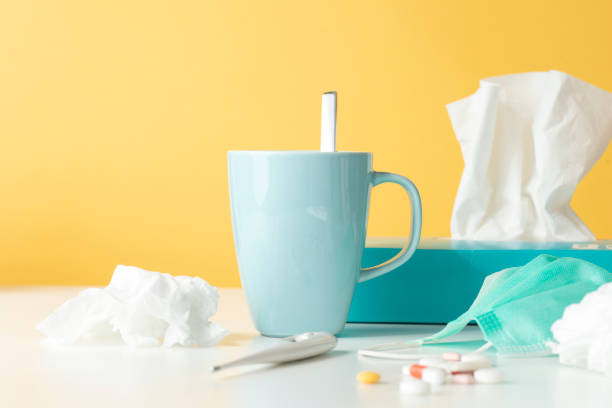 Cold and Flu Tissue box, tissues,  thermometer, protective face mask, capsules and a hot drink on white table in front of yellow wall. facial tissue stock pictures, royalty-free photos & images