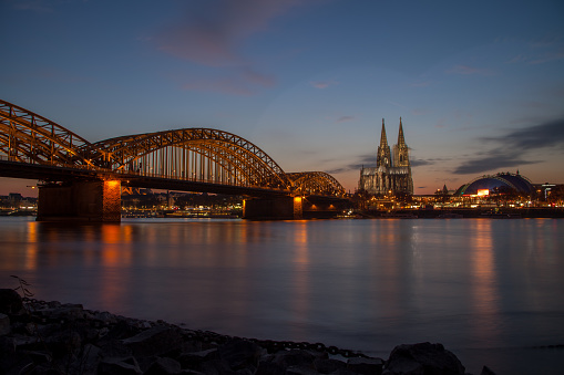 Cologne Cathedral and the Hohenzollern Bridge at twilight. Boats are parked along the river and people are passing the famous bridge.