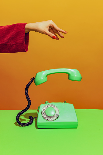 Colorful bright image of female hand holding old-fashioned green colored phone, handset falling down isolated over orange background. Concept of pop art, vintage things, mix old and modernity