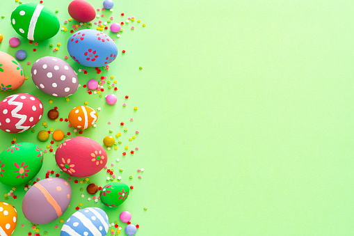 Overhead view of a group of hand painted colorful Easter eggs arranged at the left of a green background making a frame and leaving useful copy space for text and/or logo. Sugar sprinkles and candies complete the composition. High resolution 42Mp studio digital capture taken with Sony A7rII and Sony FE 90mm f2.8 macro G OSS lens
