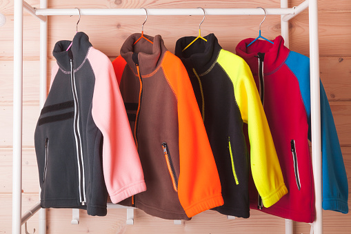 Colorful fleece jackets are hanging on hangers near wooden wall in a rack