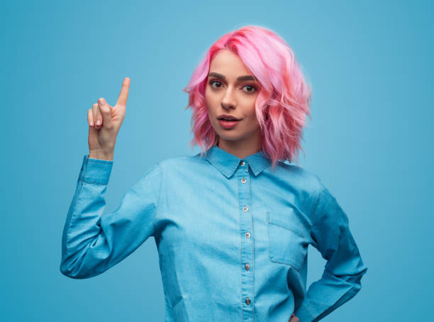 Colorful trendy woman pointing up Charming young woman with pink dyed hair wearing blue shirt and pointing up on blue background pink hair stock pictures, royalty-free photos & images